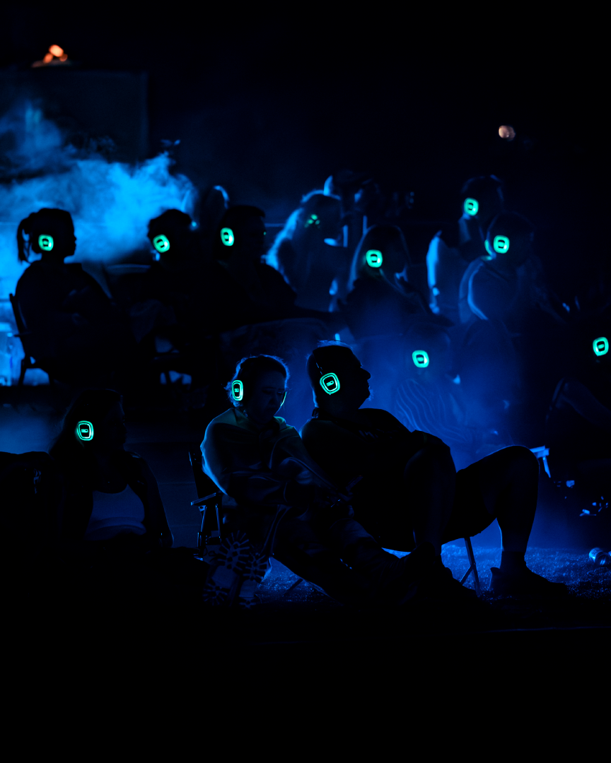 Group of people wearing illuminated headphones sitting outdoors at night, enjoying a silent disco or outdoor movie experience.