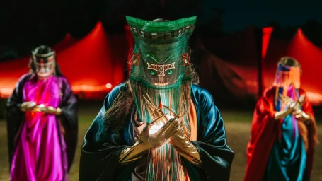 Three figures wearing elaborate ritual headwear and satin robes holding glowing lights against a dark outdoor background with red tents.