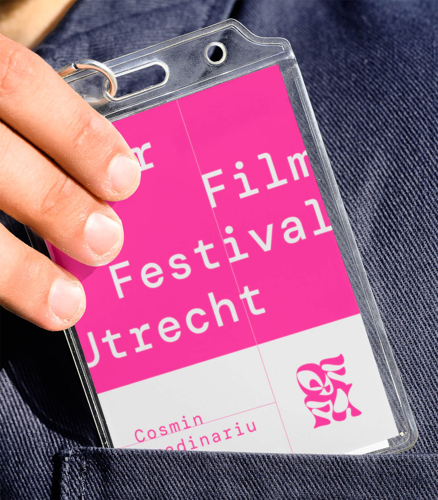 Film festival badge with pink background in pocket being held by a hand. Badge is in a transparent plastic sleeve.