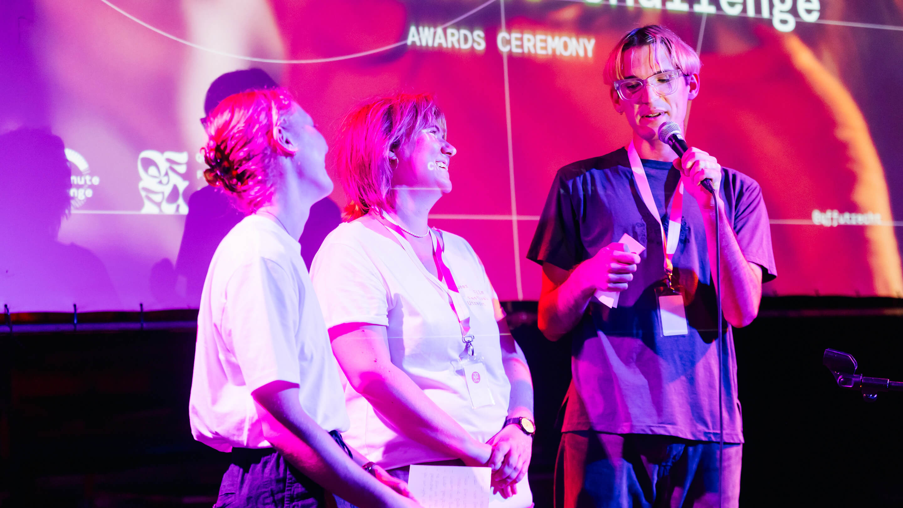 Three people on stage at awards ceremony; one male speaks into microphone in front of a projection screen in magenta lighting.
