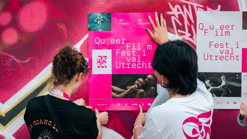 Two people attach a pink poster for the Queer Film Festival Utrecht to a graffiti-covered wall, promoting a cultural film event.