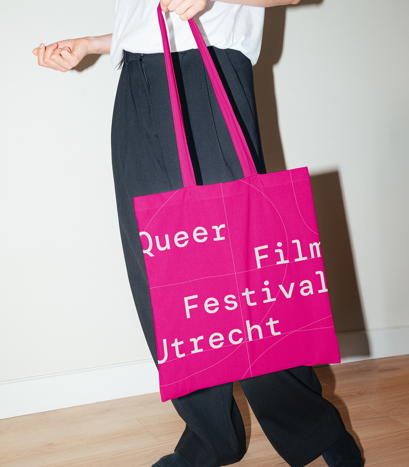 Person holding a bright pink tote bag with 'Queer Film Festival Utrecht' printed in white, paired with a white shirt and dark pants.