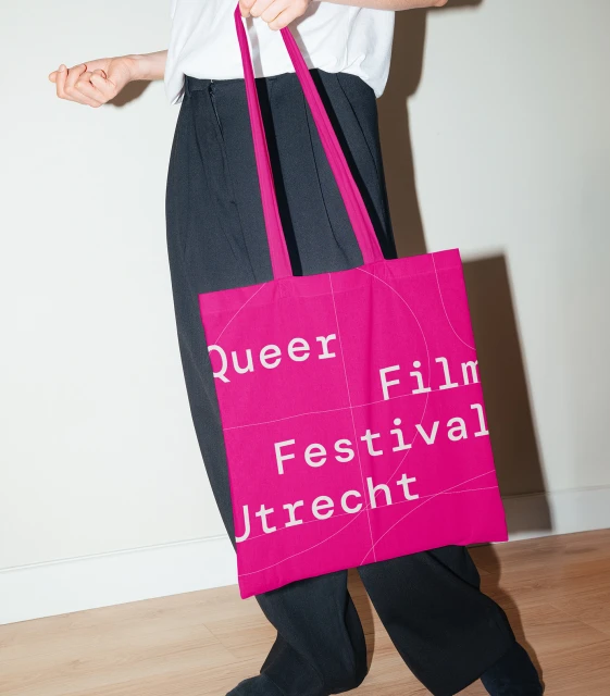 Person holding a bright pink tote bag with 'Queer Film Festival Utrecht' printed in white, paired with a white shirt and dark pants.