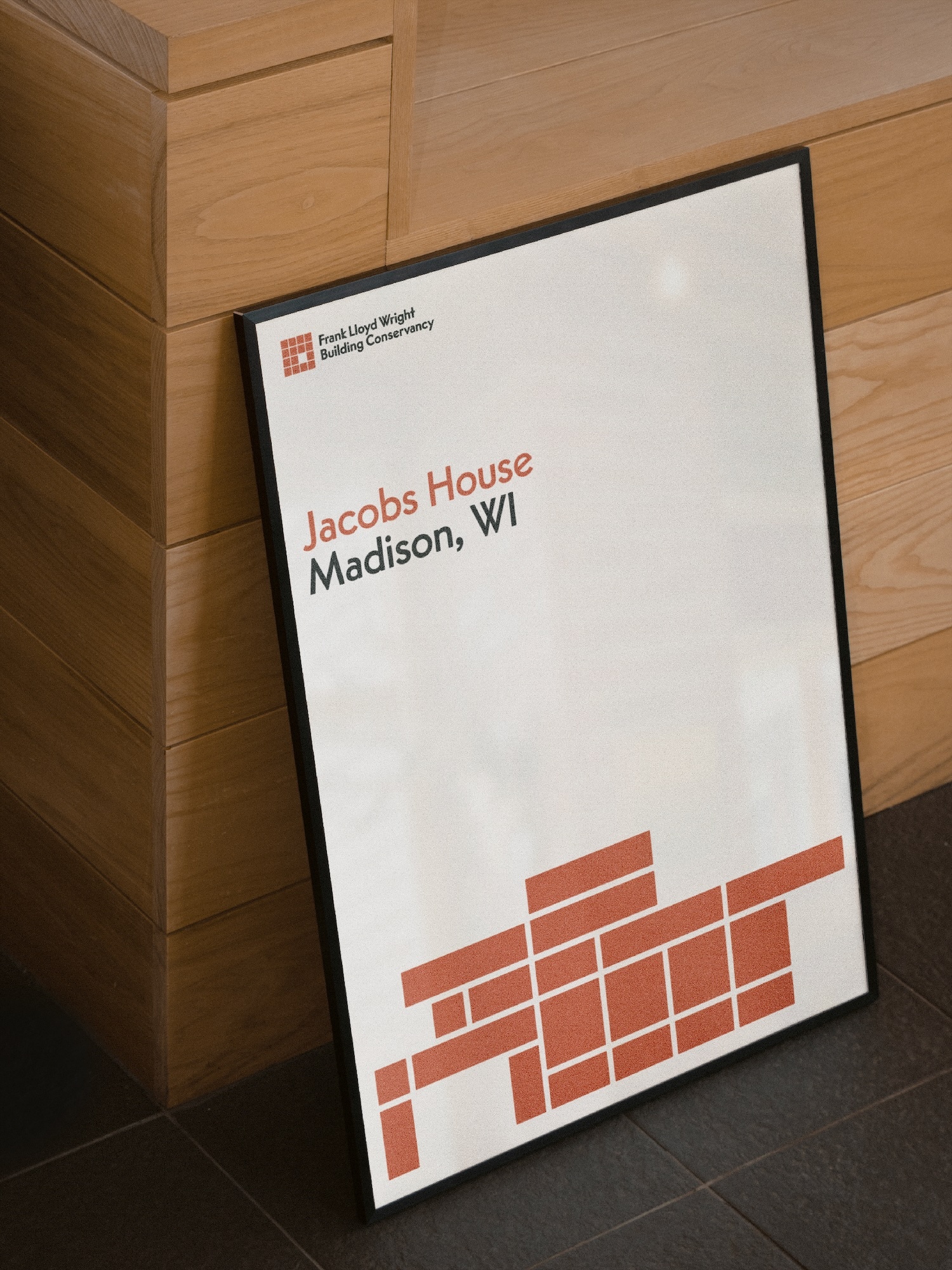 Framed poster with the Frank Lloyd Wright Building Conservancy logo and text about the Jacobs House, Madison, WI, leaning on a wall.