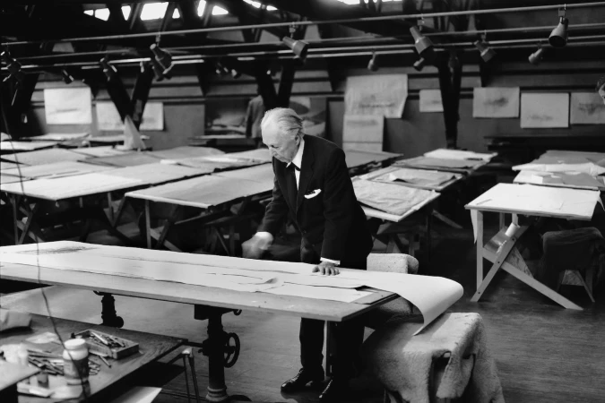 An elderly man in a suit meticulously works on a large sheet of paper, surrounded by tables and natural light in a vintage studio.