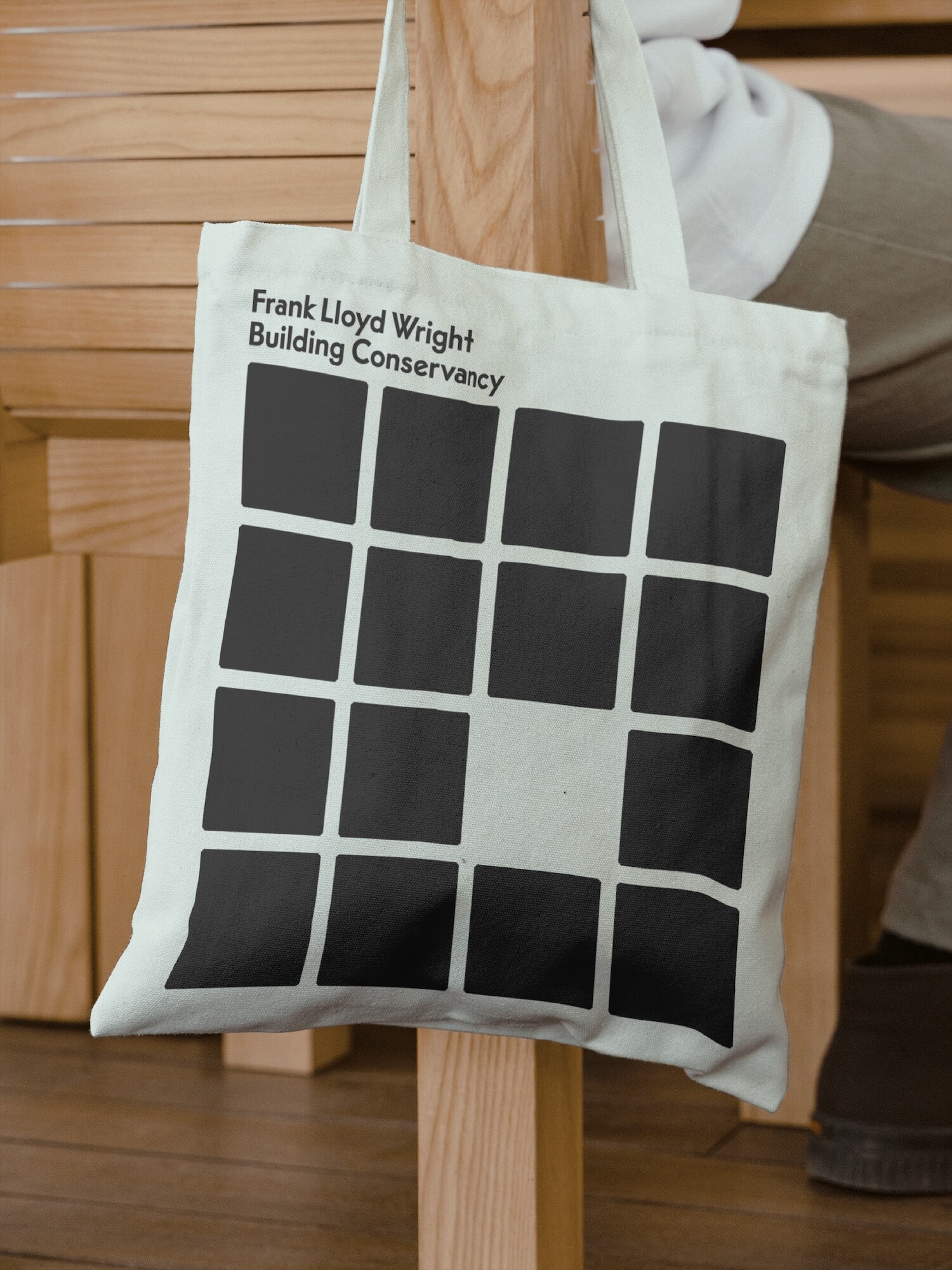 Canvas tote bag with a black geometric grid and text graphic, hanging on a wooden chair, showcasing a minimalist design.