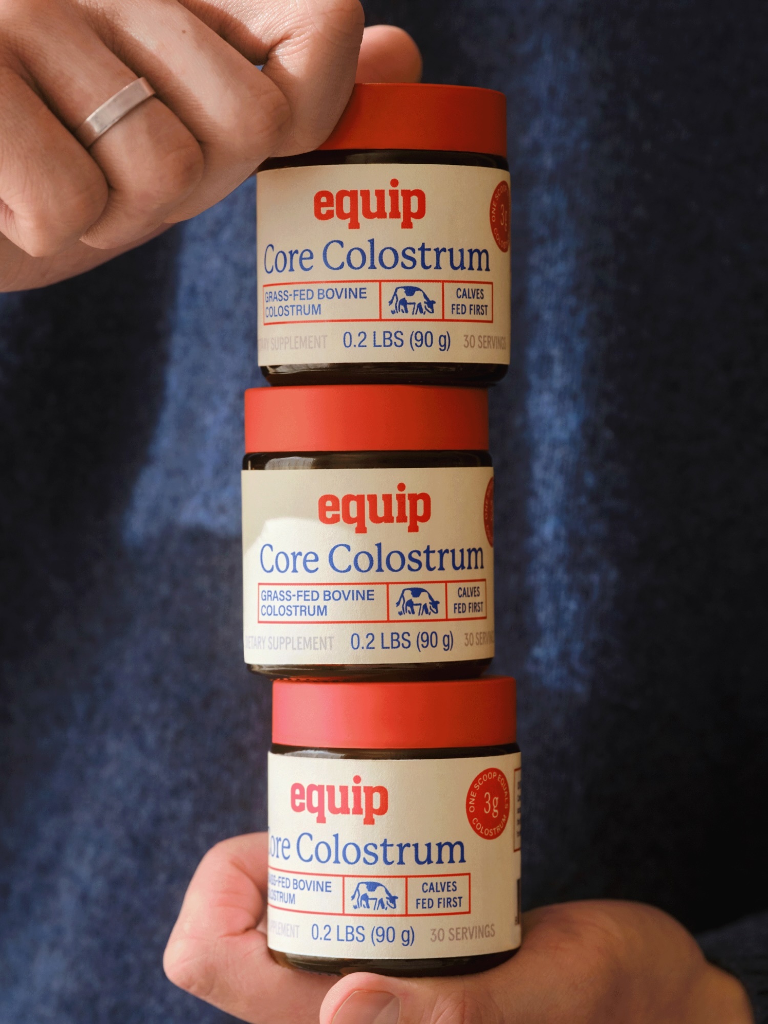Three jars of Equip Core Colostrum supplement stacked on top of each other, held by two hands, against a blurred denim background.