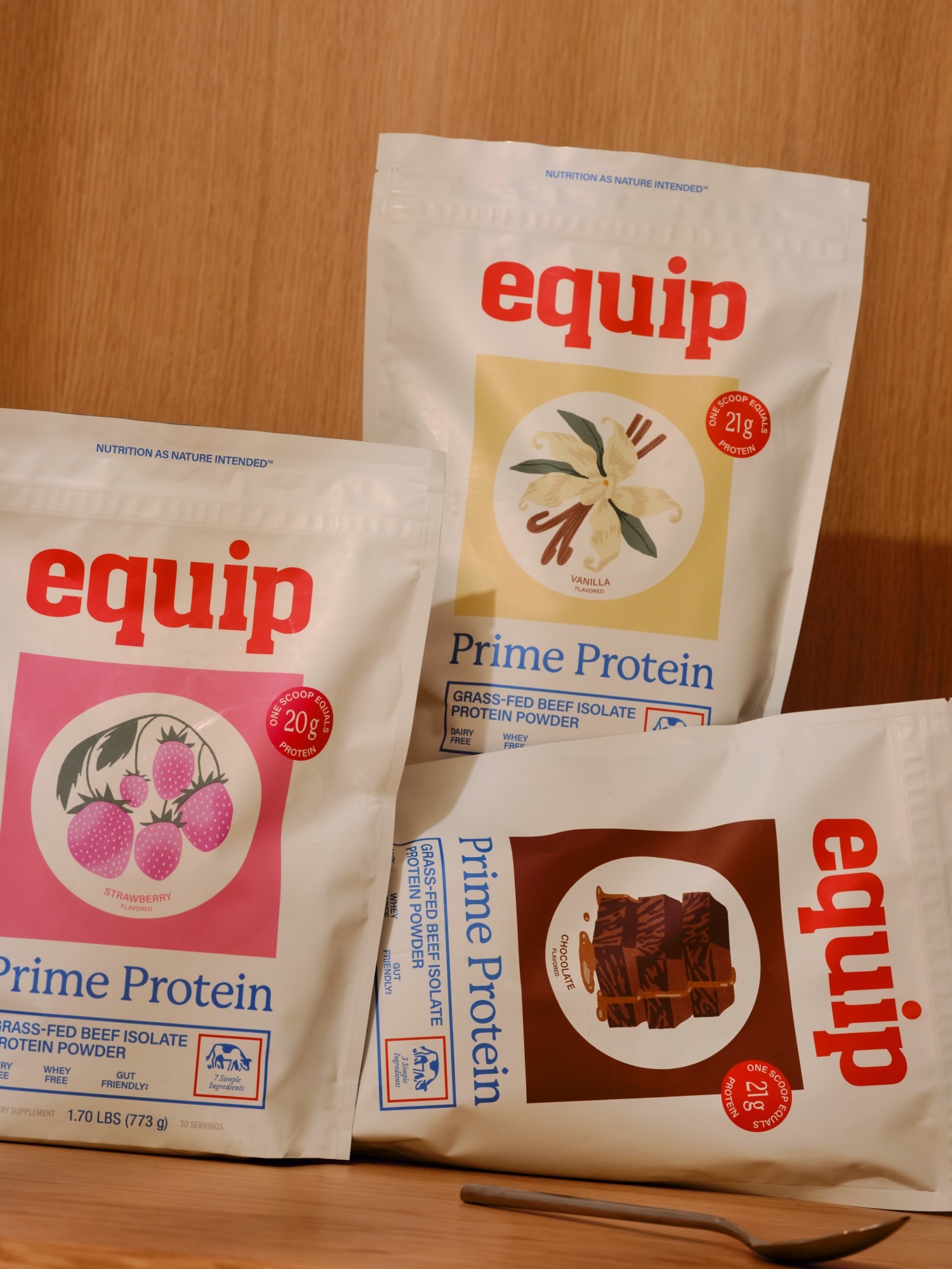 Three Equip Prime Protein bags, flavors vanilla, chocolate, and strawberry, displayed on a wooden surface with a wooden spoon.