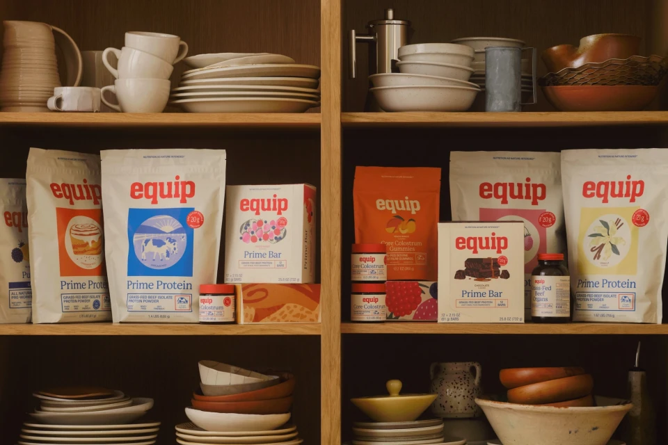 Equip brand protein powder, gummies, and bars displayed on wooden shelves with dishware in a warm, inviting kitchen scene.