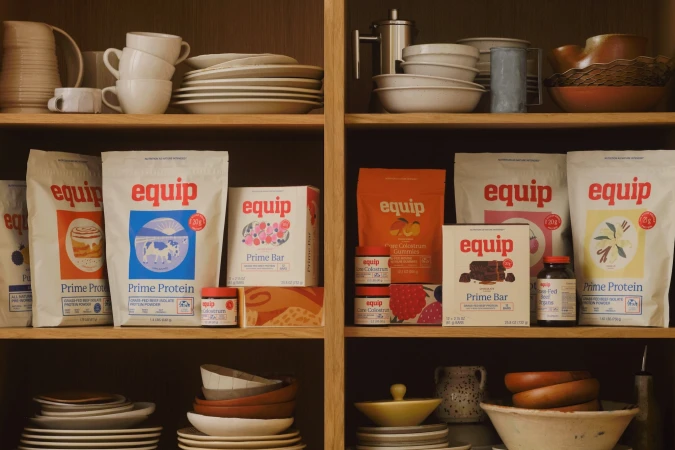 Equip brand protein powder, gummies, and bars displayed on wooden shelves with dishware in a warm, inviting kitchen scene.