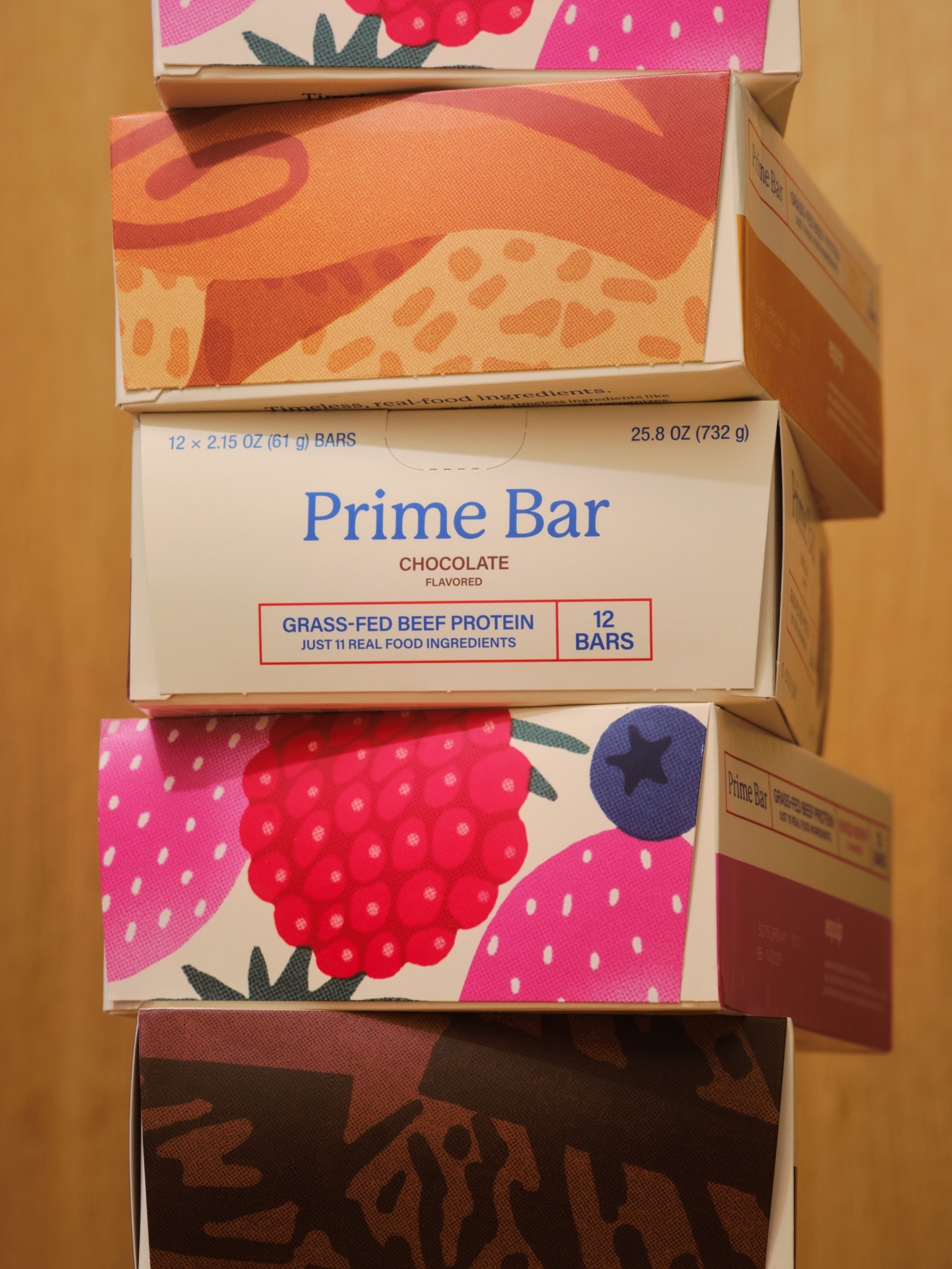 Stacked boxes of Prime Bar grass-fed beef protein bars in white boxes with fruit graphics on light wood background. 