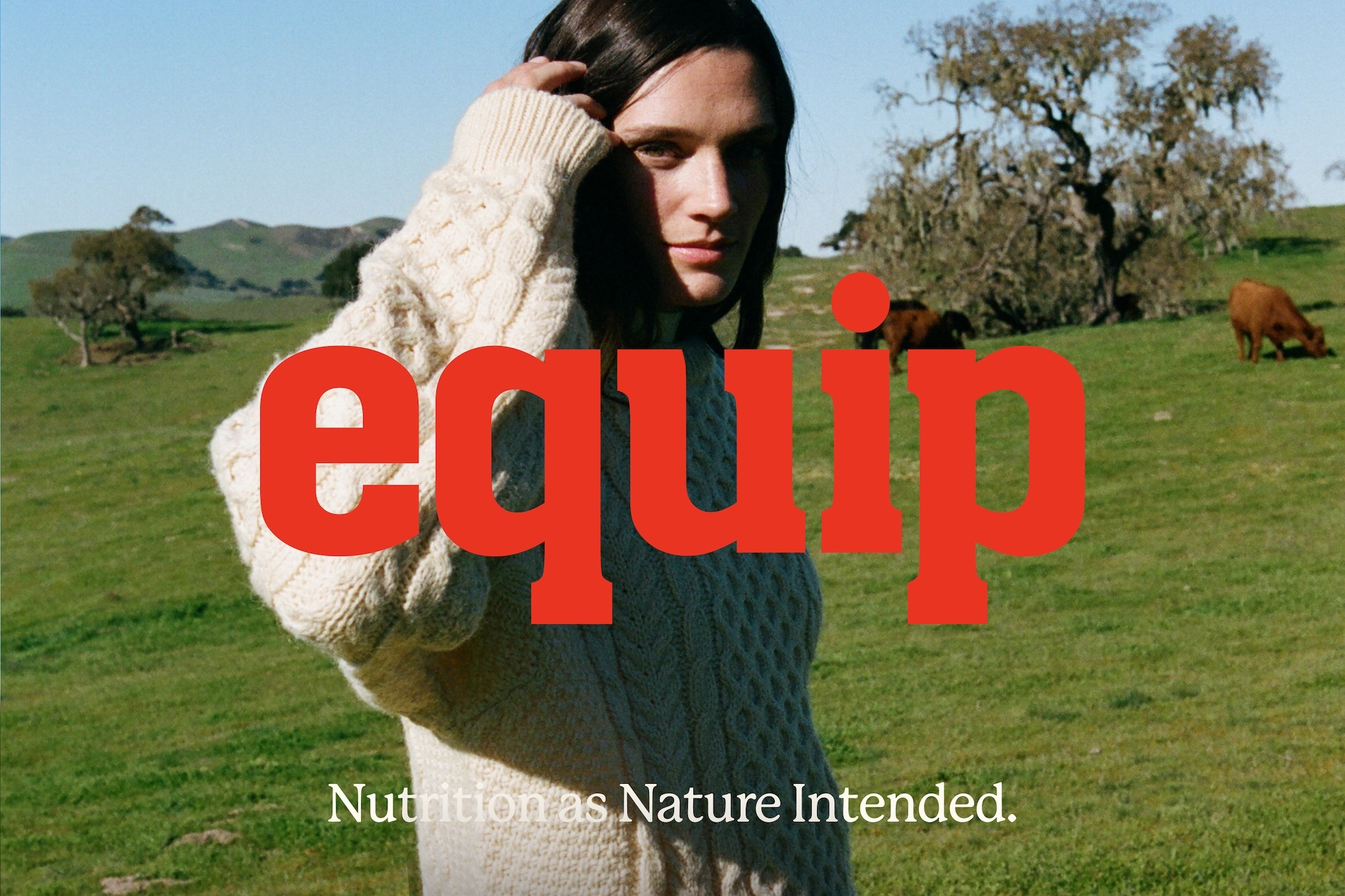 A brunette woman in a cream cable-knit sweater poses in a grassy field with grazing cows and scattered trees under a blue sky.