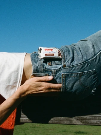 A person wearing denim overalls holds a protein bar with blue sky and green grass in the background. The bar is equip Prime Bar chocolate.