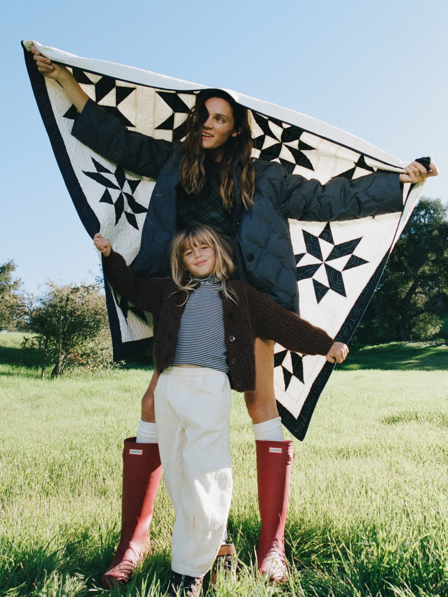 A woman and child pose in a grassy field, holding up a quilt with star patterns on it, both wearing jackets and boots in an outdoor setting.