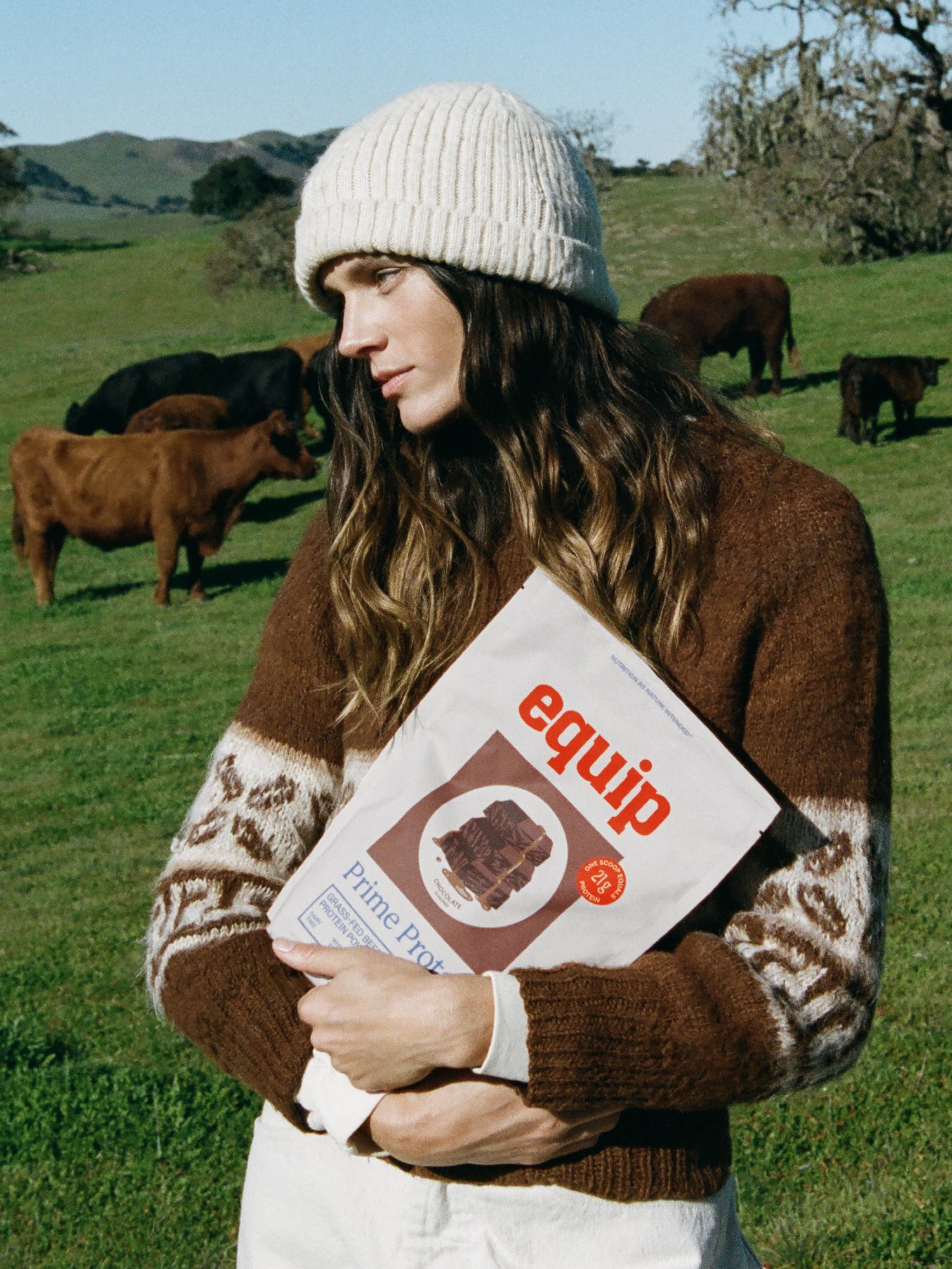 Person in a beanie and patterned sweater holds Equip Prime Protein bag in a grassy field with cows, creating a natural setting.