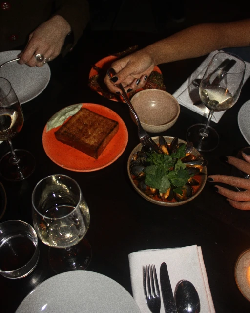 Overhead view of a dark table featuring plates of food including bread, mussels, and other dishes, along with wine glasses and cutlery.