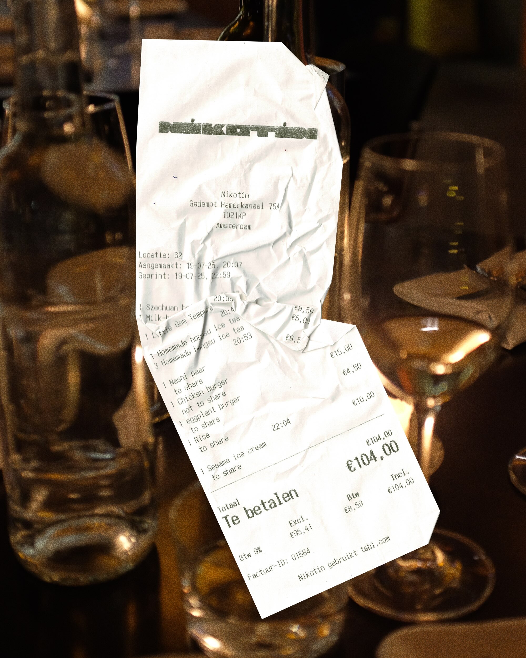 Restaurant bill on a dark table next to empty glasses. The crumpled receipt is from Nikatin in Amsterdam, totaling €104.00.