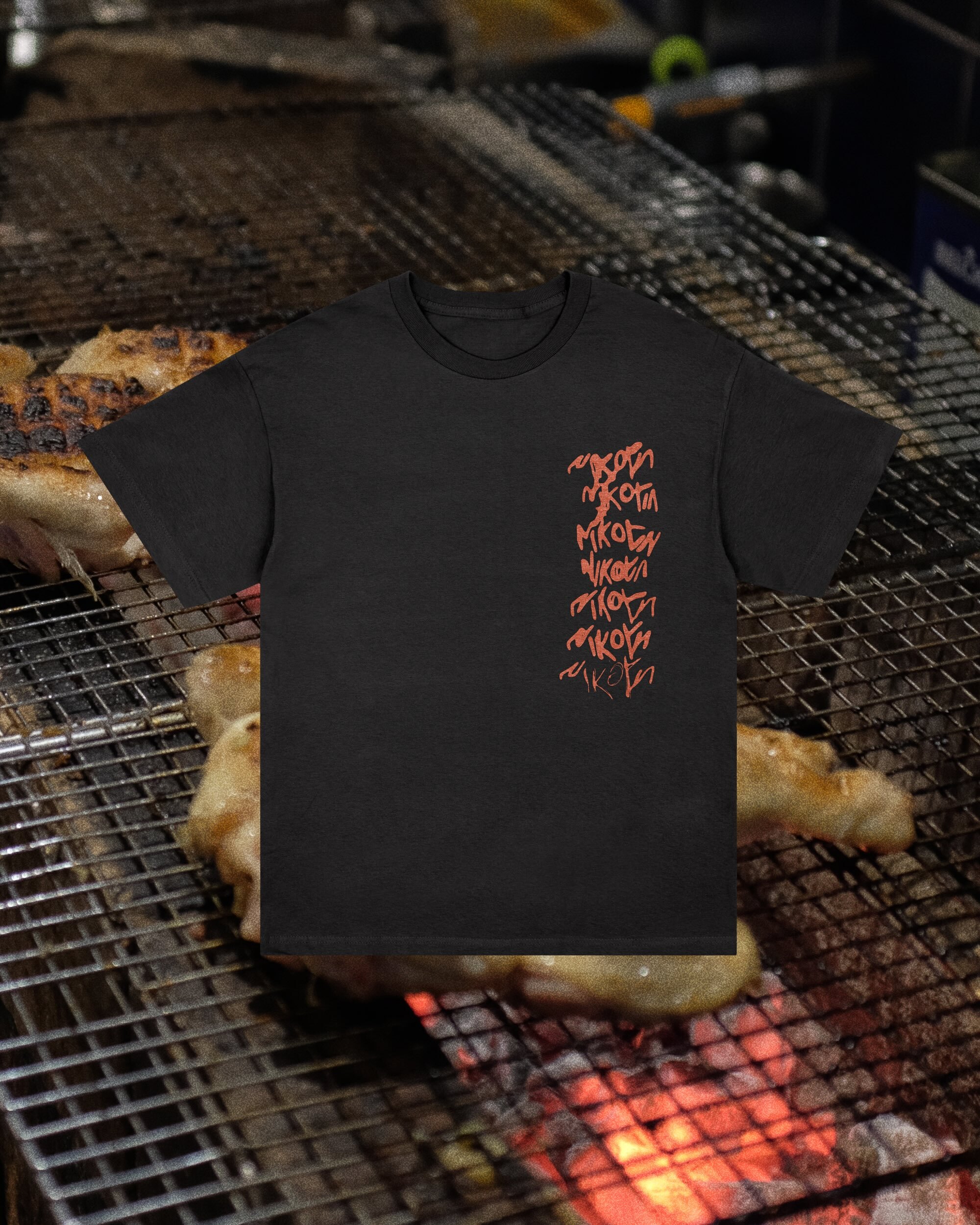 Black t-shirt with red graphics laid flat on a metal grill rack, partially covering grilling chicken in a low-angle indoor shot.