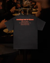 Black t-shirt featuring orange printed text on the back for Nikotin Restobar, photographed against a dim restaurant interior background.