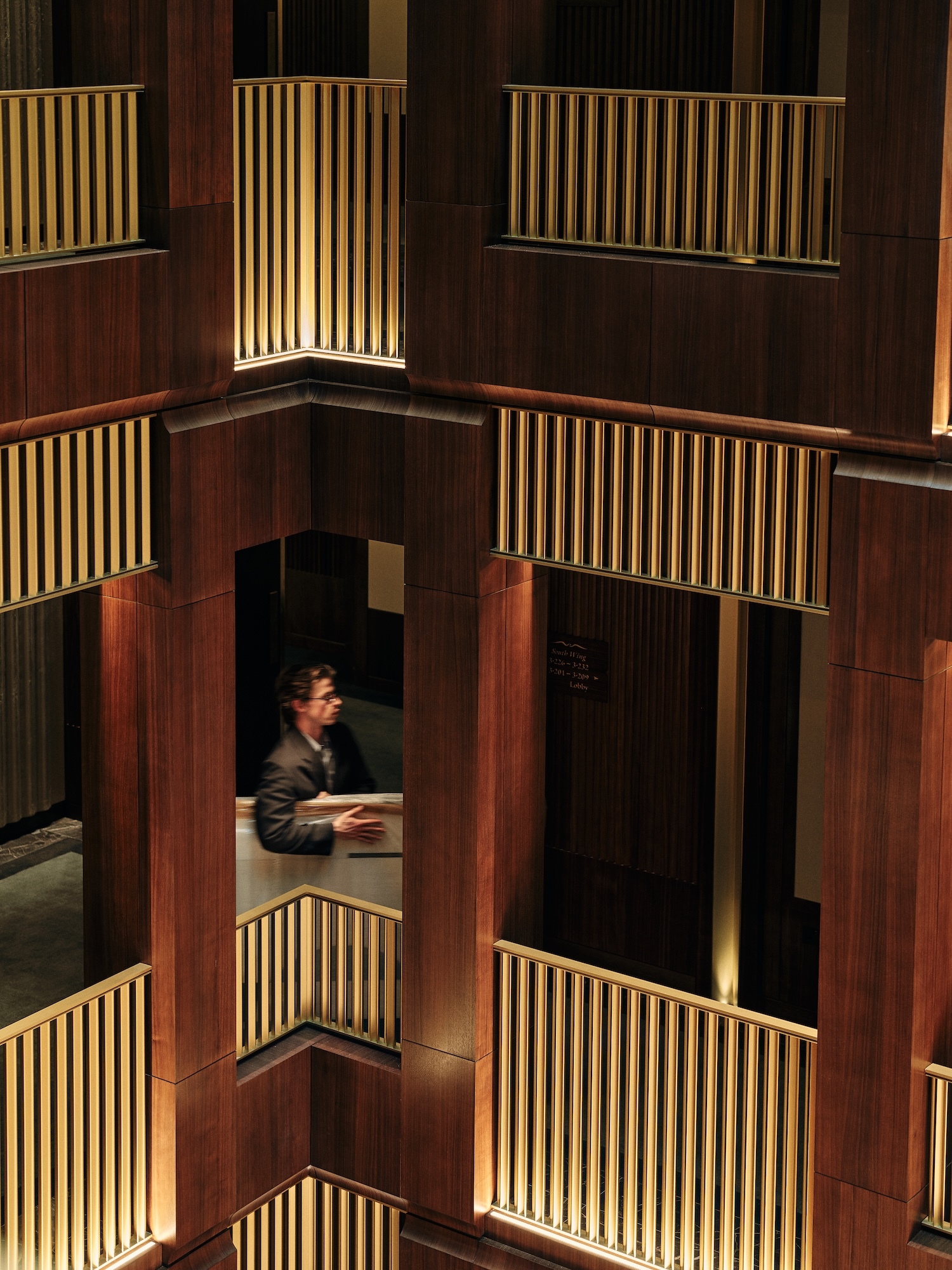 Adult male wearing a suit stands on the balcony of a wood-paneled building with gold railings. The balconies form a symmetrical pattern.