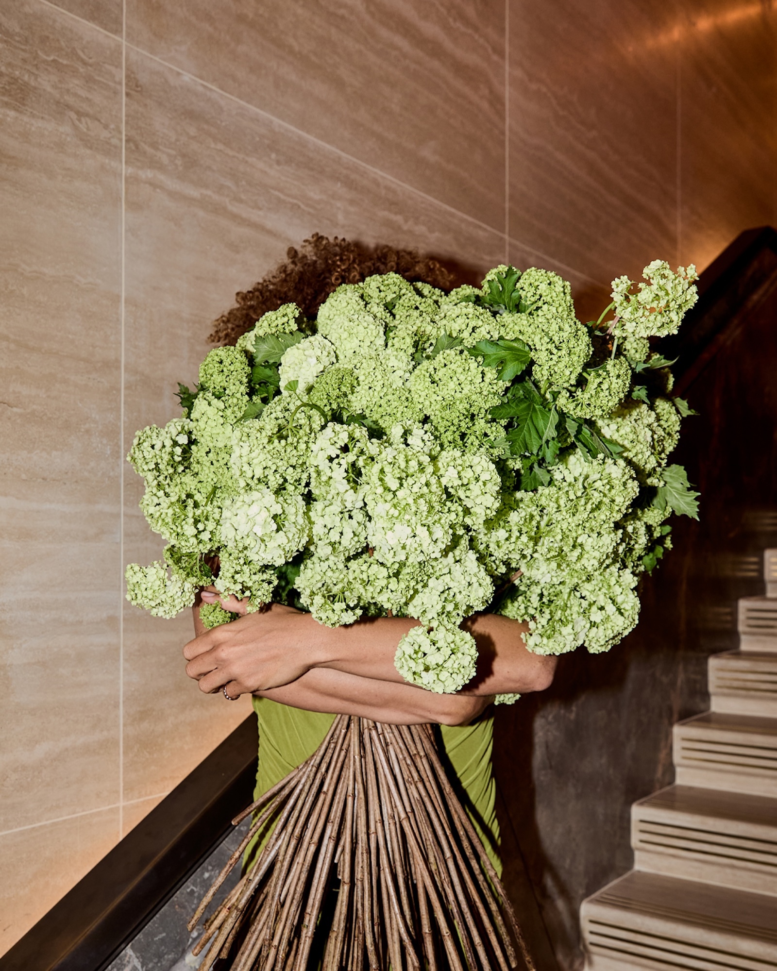 Person holding a large bouquet of green flowers indoors on a staircase. The person is wearing a green dress and the flowers obscure the face.