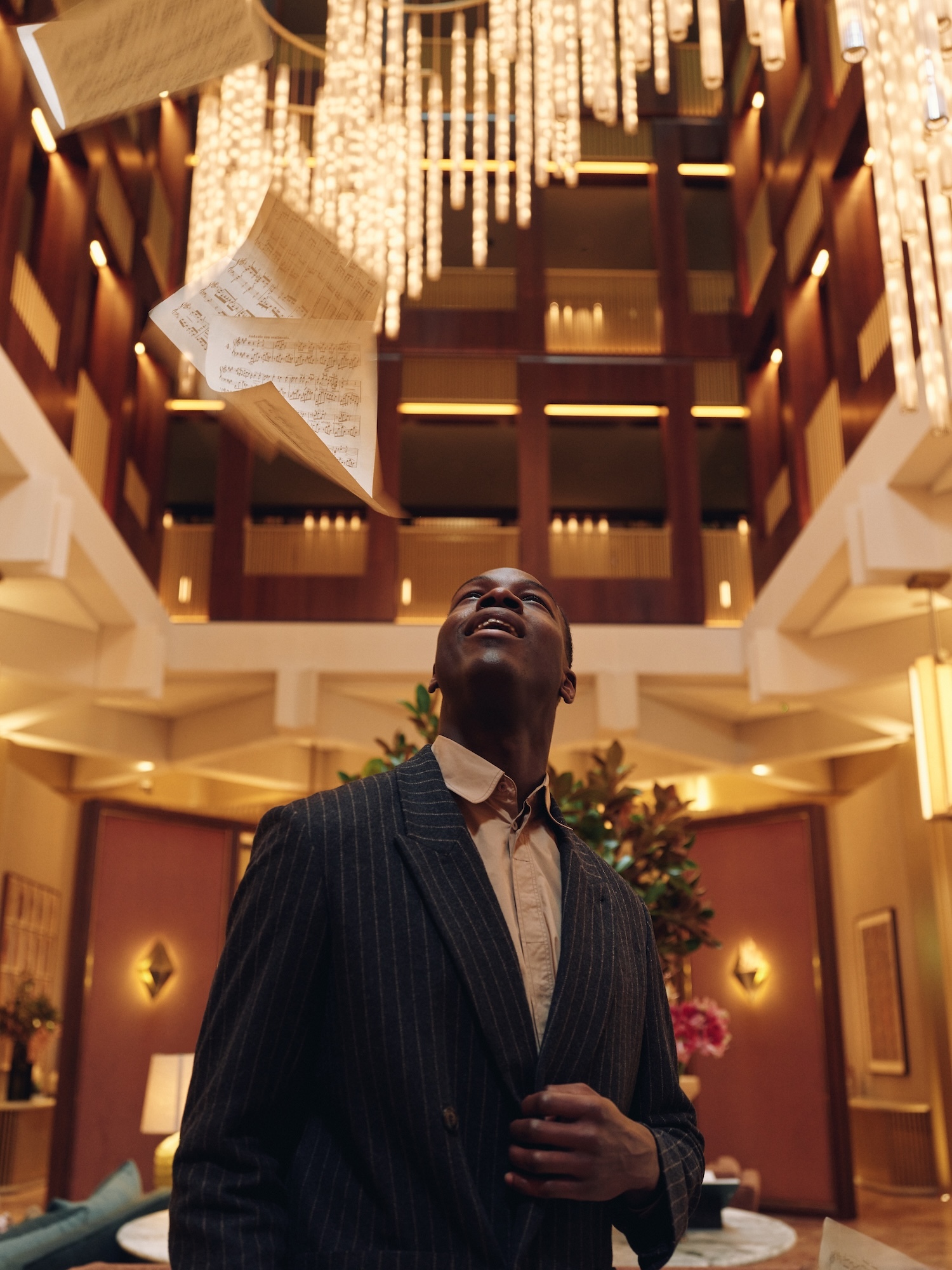 An African-American adult male in a striped suit looks up at falling sheet music in a high-ceilinged lobby with chandeliers and wood accents.