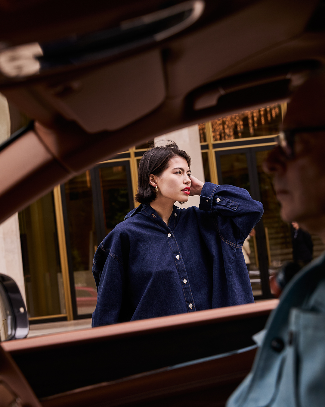 Woman in a denim shirt, viewed through a car window; second adult in passenger seat, building visible in background; car interior frame.