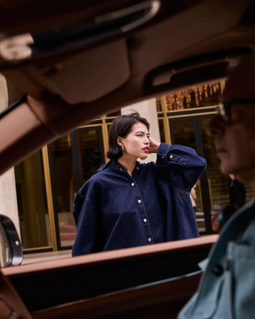 Woman in a denim shirt, viewed through a car window; second adult in passenger seat, building visible in background; car interior frame.