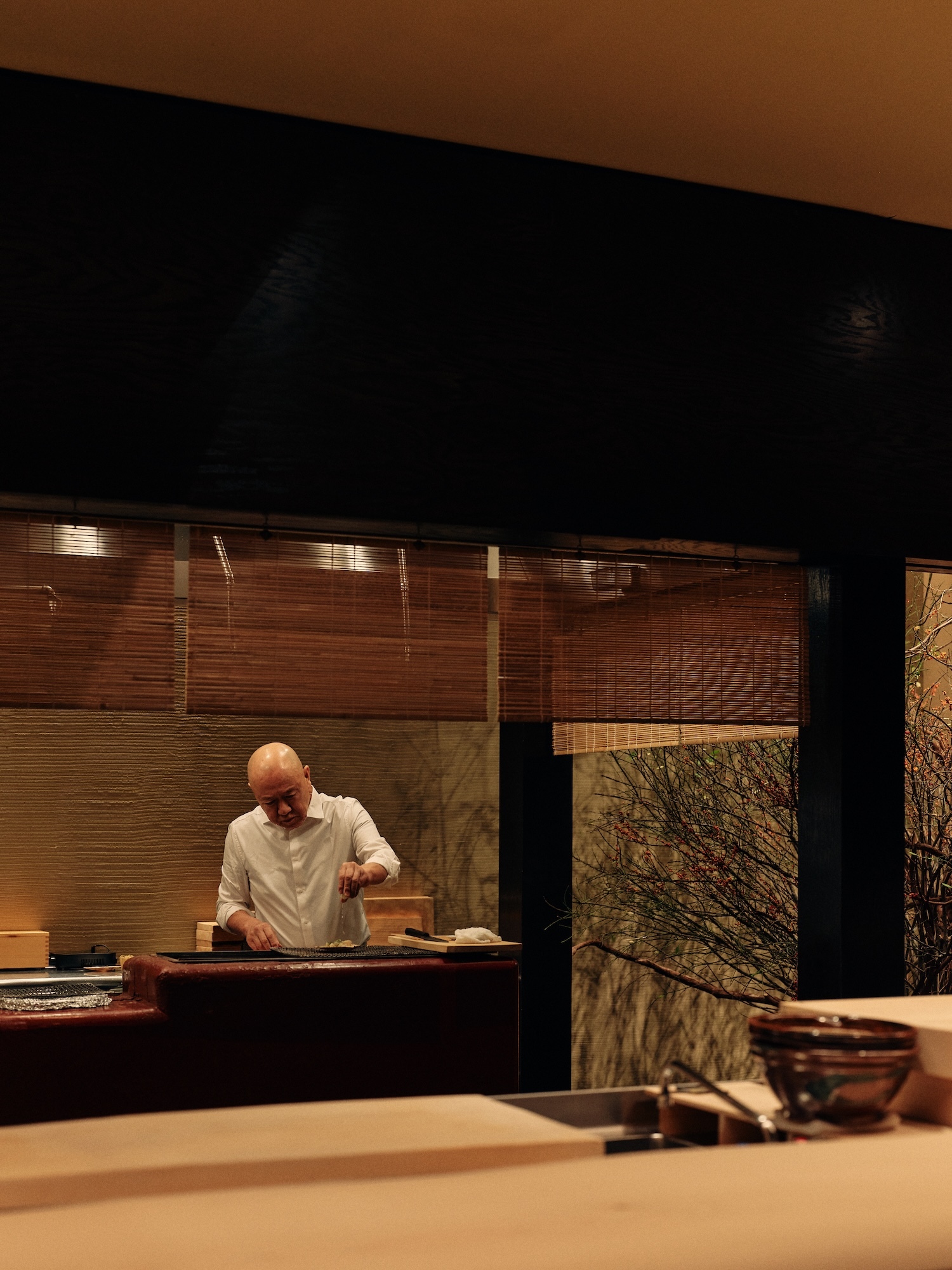 Bald adult male chef in a white shirt is preparing food at a wooden sushi counter in a warm-toned restaurant with bamboo blinds.