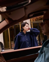 Woman in dark denim shirt standing outside near a building, viewed through the open window of a car.