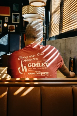Back view of a seated adult male wearing a t-shirt with Gimlet text and logo, at a table with sunlight and shadows in a dark interior.