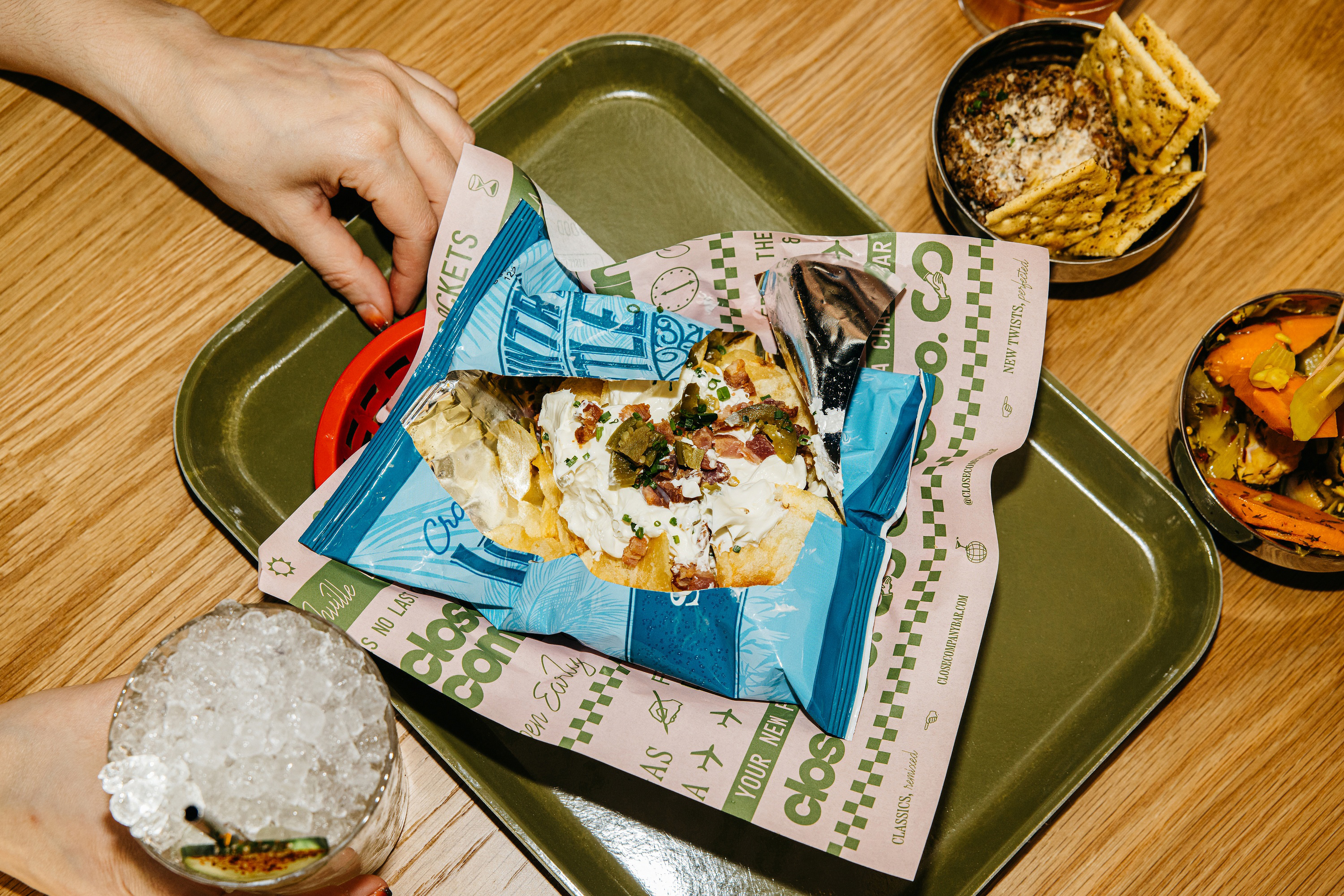 Overhead shot of chip nachos served inside of a chip bag, set on a green tray, beside a beverage and metal bowls on a wooden table.