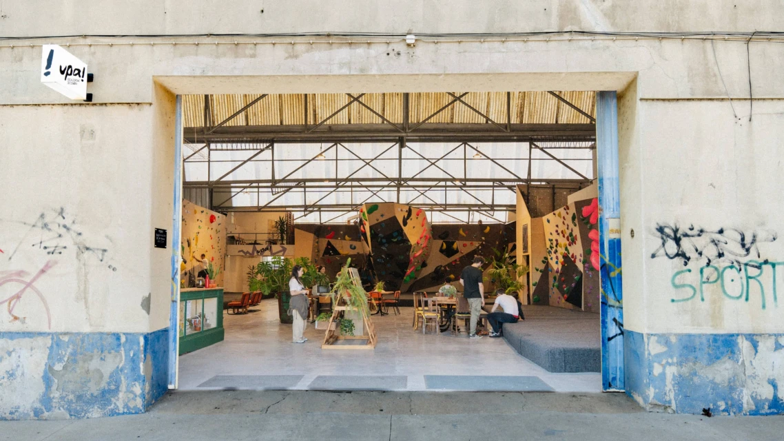 Indoor rock climbing gym featuring people, climbing walls with colorful holds, and an open doorway to the exterior.