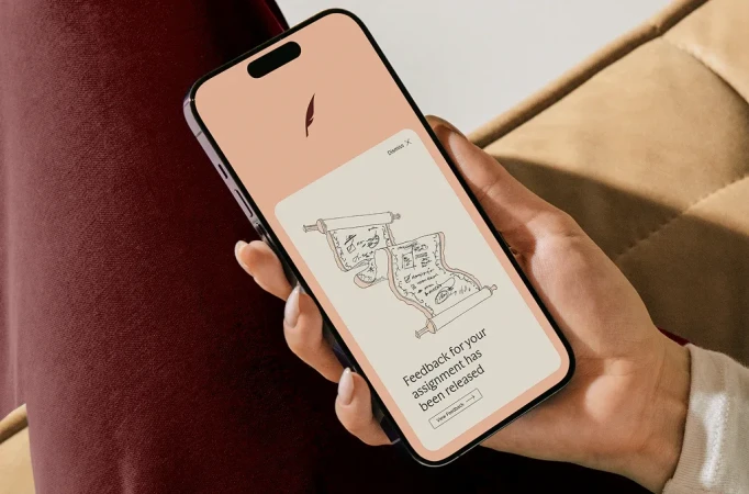 Close-up of a person holding a smartphone displaying an assignment feedback notification with a scroll illustration on a peach background.