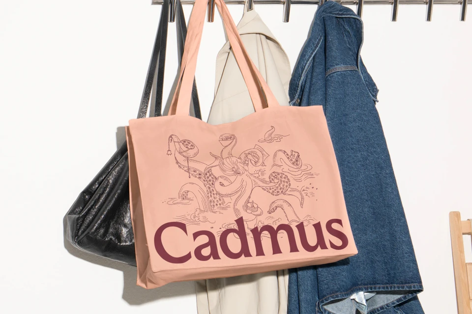 Close-up of a light orange tote bag with an octopus illustration and the text “Cadmus” hanging from a coat rack alongside other bags and clothes.