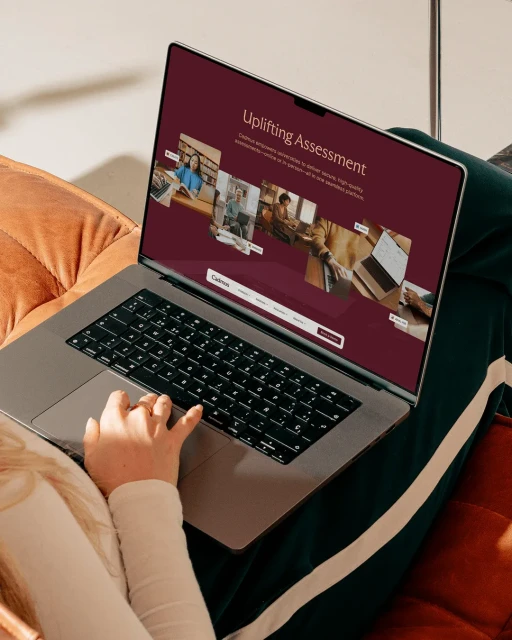 Woman using a laptop displaying a website with the text 'Uplifting Assessment'. Laptop sits on a cushion on fabric. Indoor lighting.