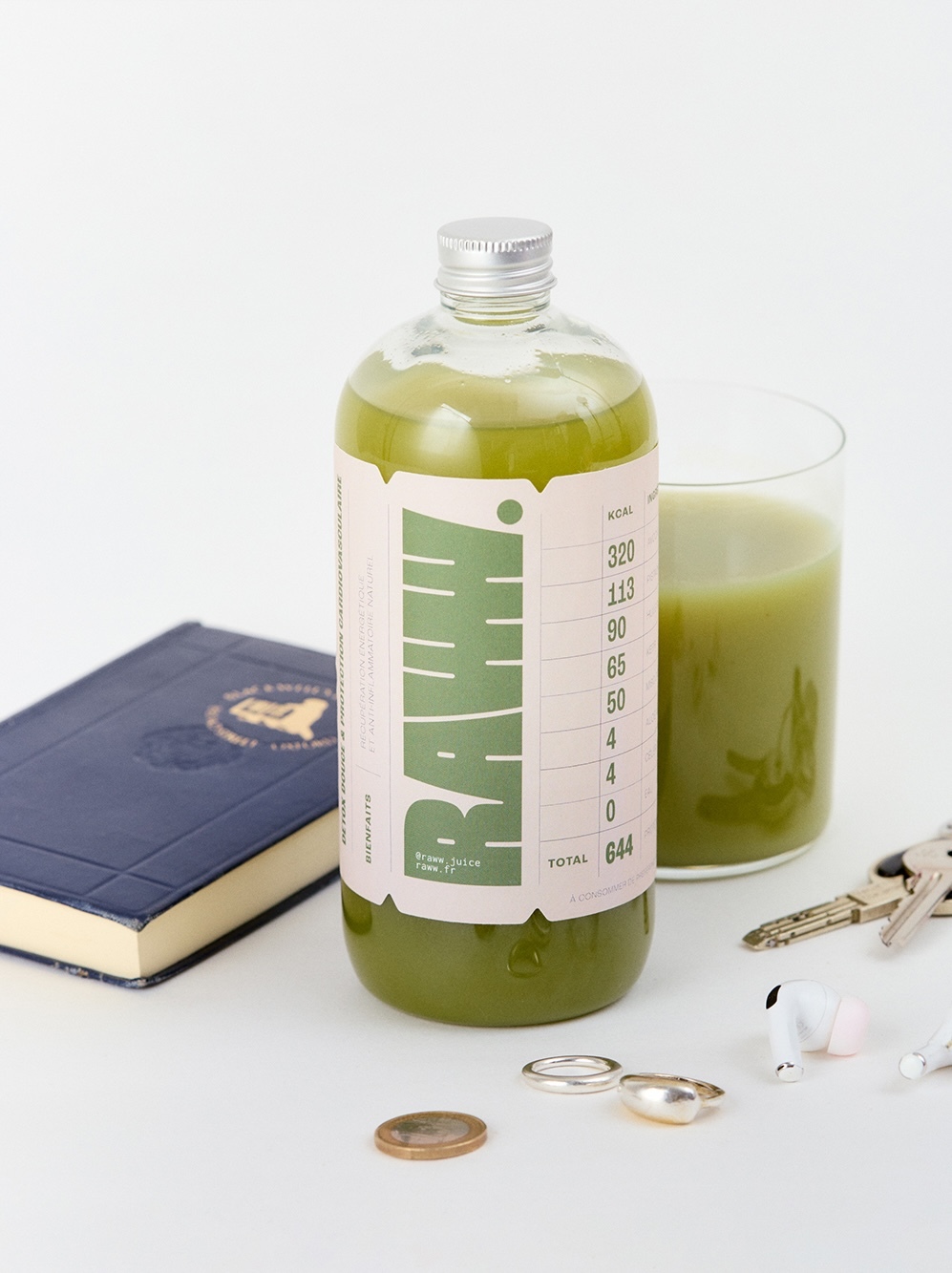 Bottle of green juice next to glass of juice, diary, coin, rings, keys, and earbuds on a white surface in a studio setting.