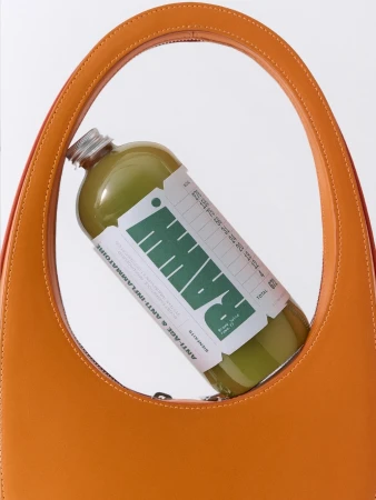 Orange leather handbag showcases a glass bottle of green juice with a label, presented against a clean, white background.