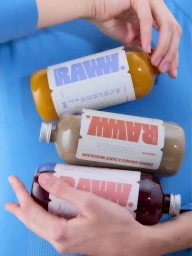 Hands holding three glass bottles of Raww juice against a solid blue background