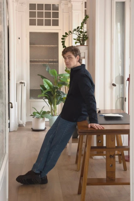 A fair-skinned young adult male with curly light brown hair wearing a dark jacket, blue jeans, and black shoes leans against a wooden desk in an indoor setting.