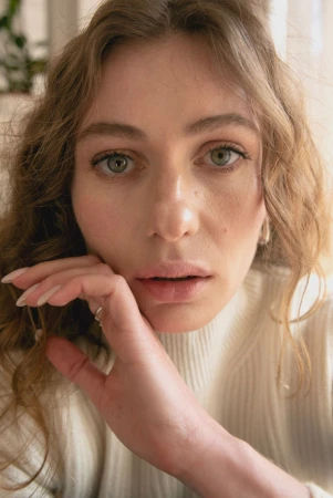 Close-up portrait of a woman with curly hair and a white turtleneck sweater resting her hand near her face.
