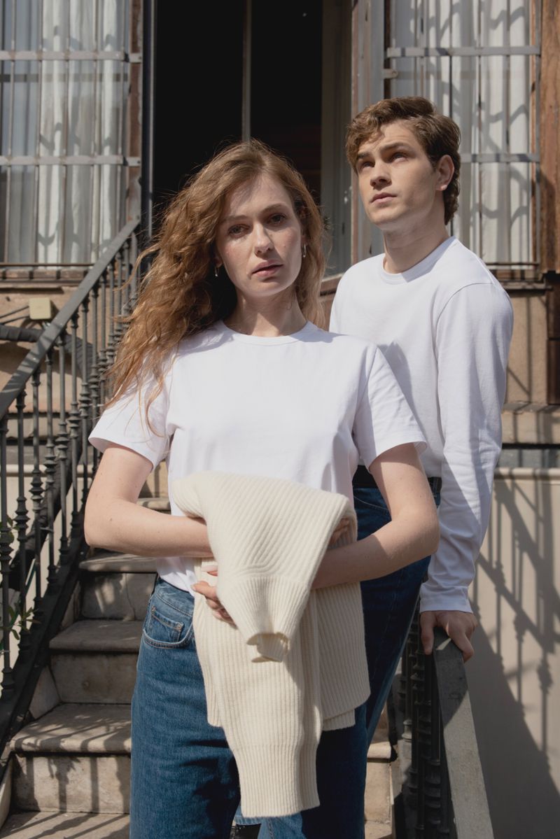 A young couple poses in front of a building with stairs, wearing white T-shirts and jeans, holding a sweater on a sunny day.