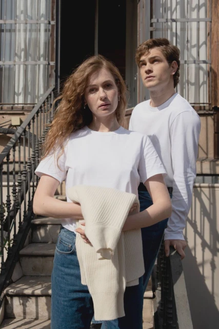 A young couple poses in front of a building with stairs, wearing white T-shirts and jeans, holding a sweater on a sunny day.