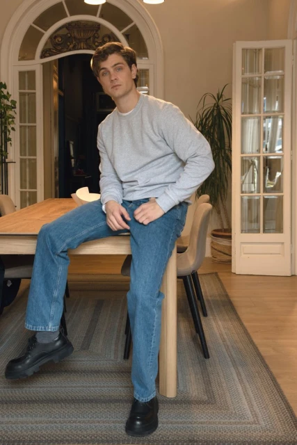 Adult man in casual clothing sits on a wooden table indoors, with chairs and a rug visible in the background.