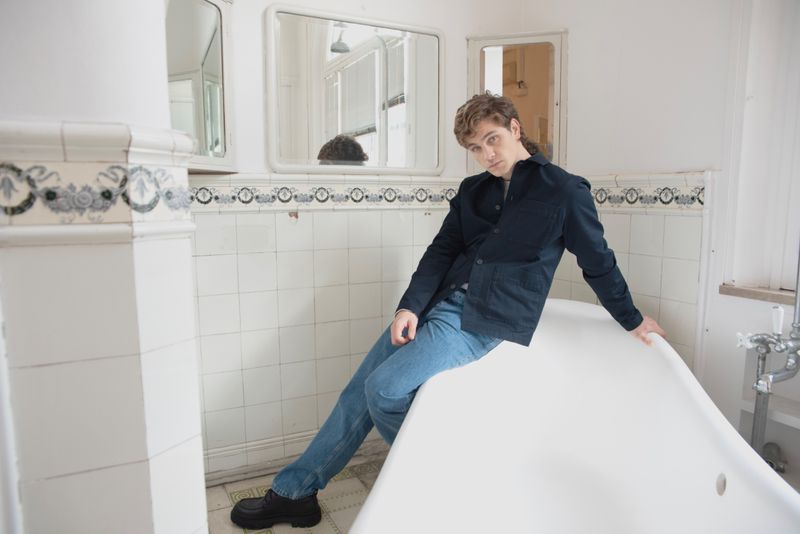 A young man sits on the edge of a vintage white bathtub in a tiled bathroom wearing jeans and a black jacket, looking at the camera.