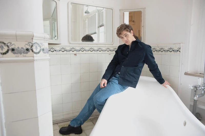 A young man sits on the edge of a vintage white bathtub in a tiled bathroom wearing jeans and a black jacket, looking at the camera.