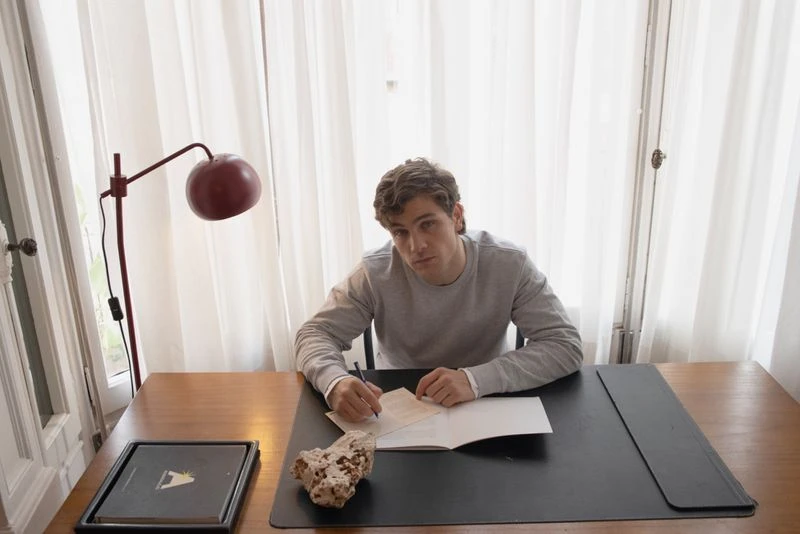 Man with curly hair sits at a wood desk by a window writing in a notebook, illuminated by a red lamp. Home interior.