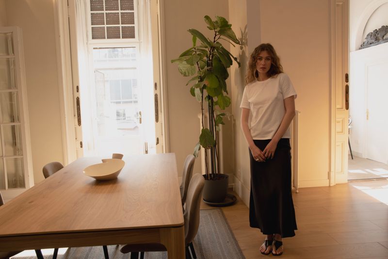 Indoor shot of a woman in a white t-shirt and black skirt standing in a well-lit room with a dining table and potted plant.