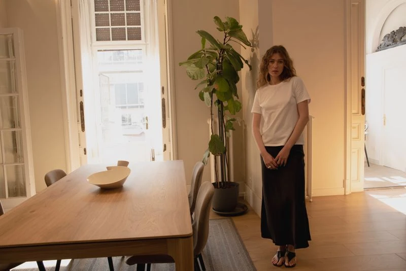 Indoor shot of a woman in a white t-shirt and black skirt standing in a well-lit room with a dining table and potted plant.