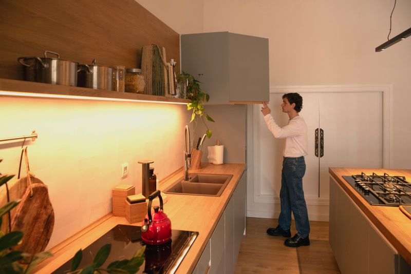 Young man in a white shirt stands in a brightly lit kitchen, reaching for a gray cabinet above the sink and stove.